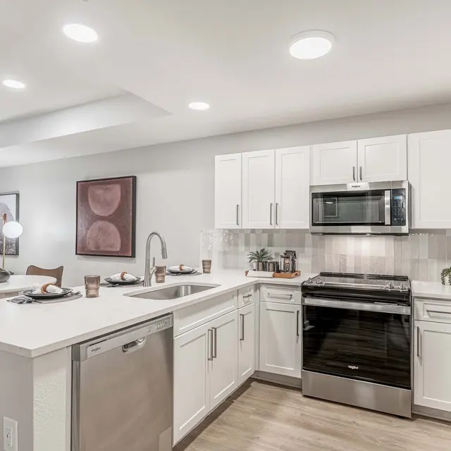 A modern kitchen featuring white cabinetry, stainless steel appliances, and a dishwasher. The kitchen island is adorned with decorative plates and has a sink. There is a living area visible in the background with a sofa and artwork on the wall.