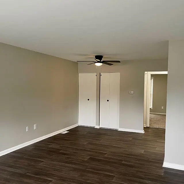A spacious empty living room featuring light gray walls, dark wood flooring, and a ceiling fan. There are two closed white doors on one side, leading to closets or storage, and an open doorway on the right side leading to another room.