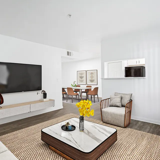 A stylish modern living room featuring a large flat-screen TV mounted on the wall, a coffee table with decorative items, and cozy seating. A dining area can be seen in the background.