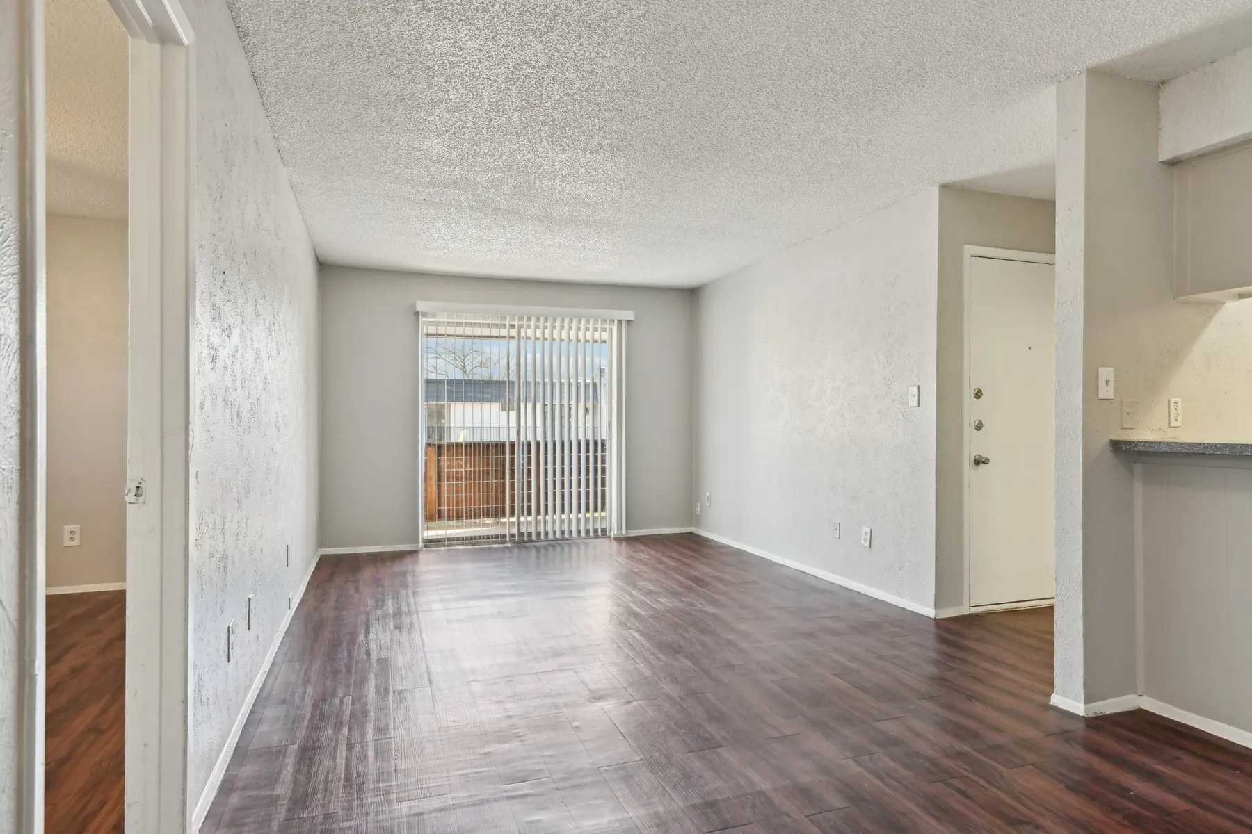 A spacious, empty room with hardwood flooring, featuring a large window with blinds letting in natural light and a door leading outside. Walls are painted in a light shade, and there is an entrance to another room visible in the background.