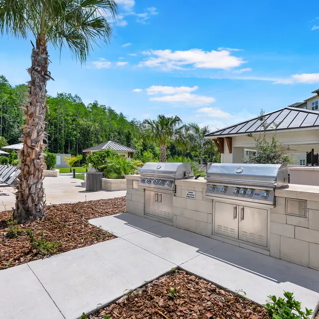 Outdoor Grilling Area A modern outdoor grilling area featuring stainless steel barbecue grills, surrounded by green landscaping and palm trees, with lounge chairs and a pool area in the background under a bright blue sky.