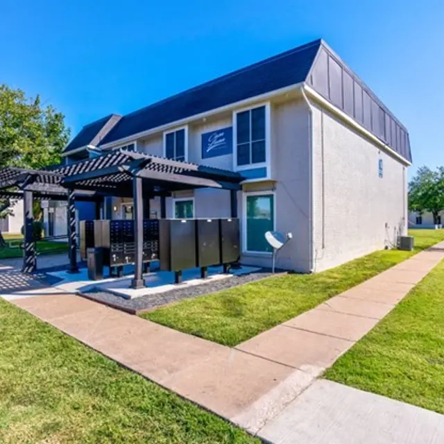 Exterior view of an apartment complex with a landscaped lawn and a shaded patio area.