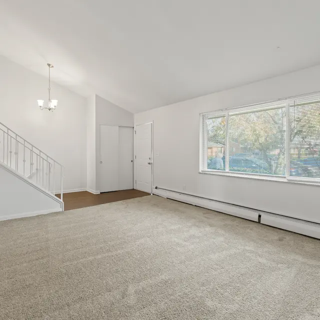 A spacious living room with light-colored carpet and white walls. There is a large window allowing natural light, and a staircase leading to an upper level. A chandelier hangs from the ceiling.