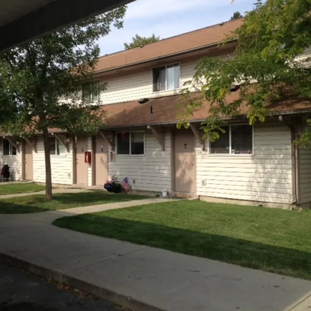 Apartment Complex Overview A row of two-story apartment buildings with a grassy lawn in front, surrounded by trees. The buildings have beige siding and brown roofs, with some visible windows and doors.