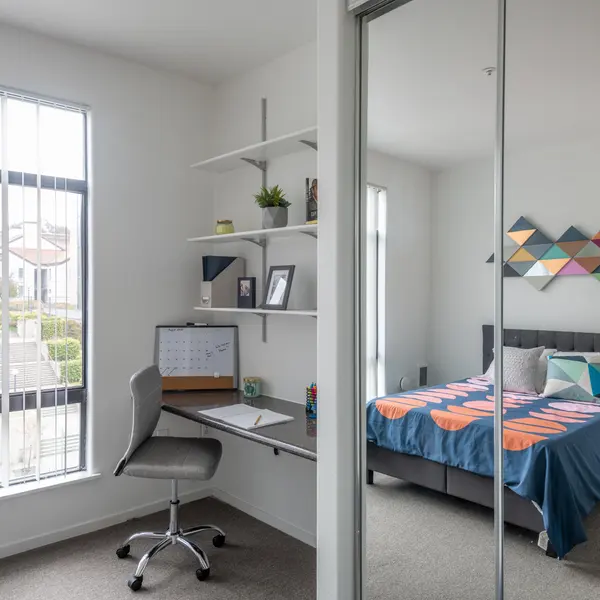 A modern bedroom featuring a symmetrical design. On the left, there's a desk with a computer setup, a calendar, and open shelving above it. On the right side, a neatly made bed with a blue and orange patterned blanket and various pillows. Large windows allow natural light to brighten the space.