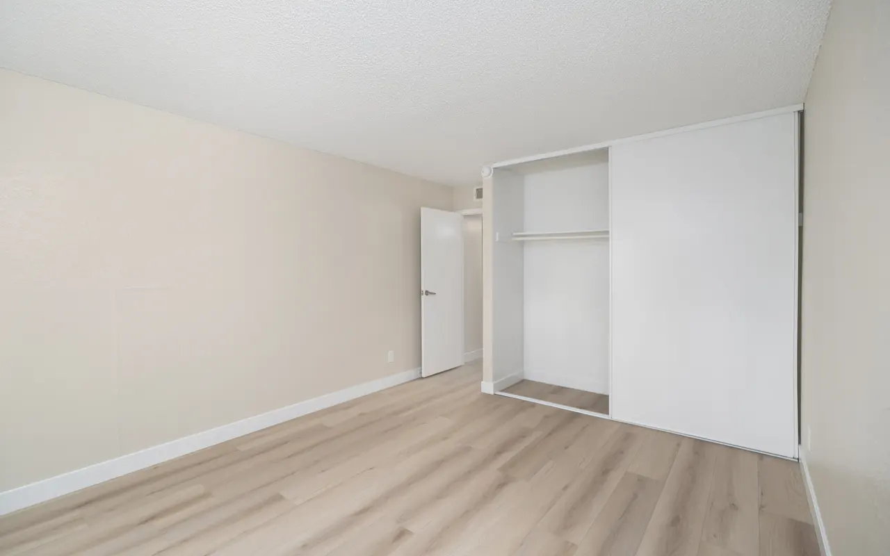 A spacious and empty room with light-colored walls and a wooden floor, featuring a closet with sliding doors and a white door leading to another area.