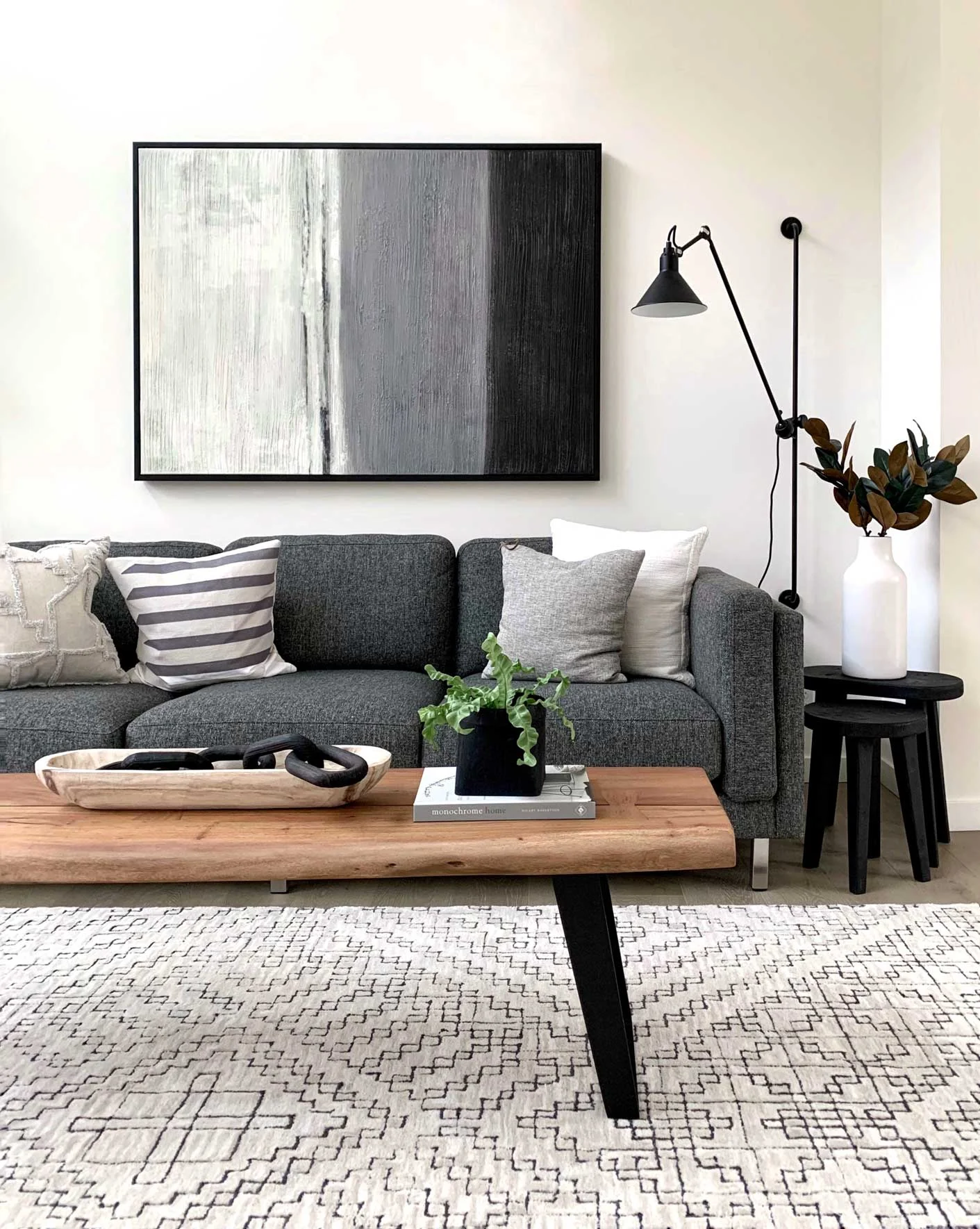 A modern living room featuring a grey couch with several cushions, a wooden coffee table with decorative items, a floor lamp, and a minimalist artwork on the wall.