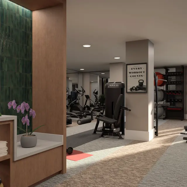 A spacious fitness center with equipment and natural light. A wooden shelf displays folded towels and a vase of orchids. There are various workout machines and free weights visible in the background.