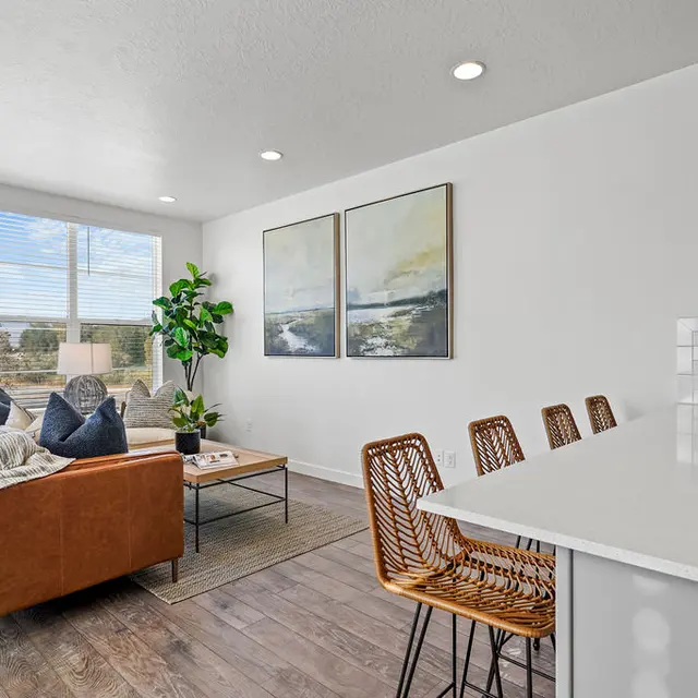 A modern living room featuring a brown leather sofa, a coffee table, and decorative plants, with large windows allowing natural light. There are two framed abstract artworks on the wall and barstools at a countertop in the background.