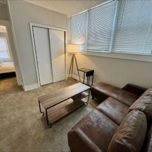 A small, cozy living area featuring a brown leather couch, a wooden coffee table, and a standing lamp, with a doorway leading to a room with a bed in the background.