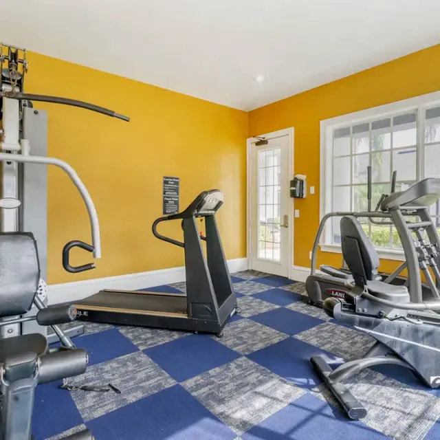 Interior view of a gym with exercise equipment including a multi-station machine, a treadmill, and an elliptical machine against a bright yellow wall.