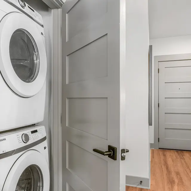 A laundry area with stacked white washer and dryer units, seen through an open gray door that reveals a hardwood floor and a glimpse of a hallway and another door.