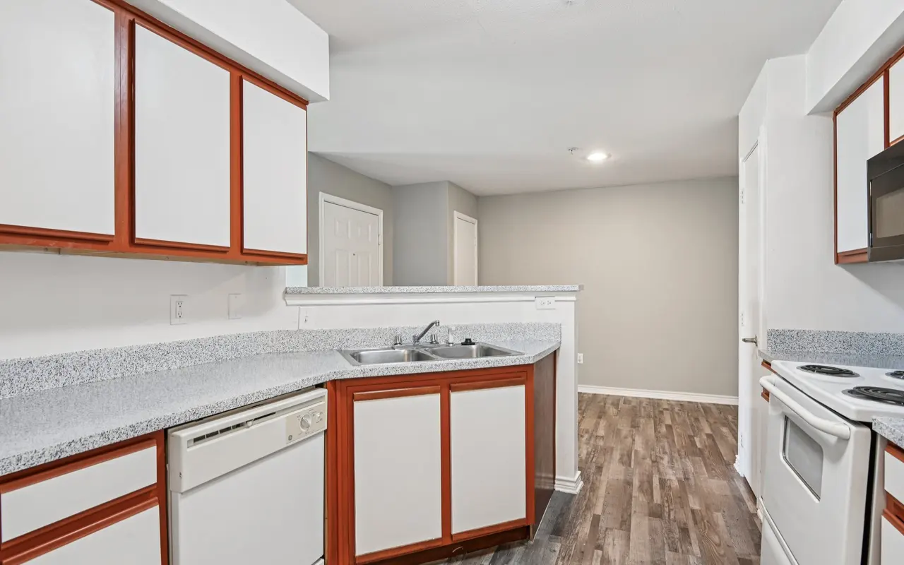 A modern kitchen with white cabinetry and contrasting red accents. It features a countertop with a sink, a dishwasher, and an oven. The flooring is a brown wood-like laminate.