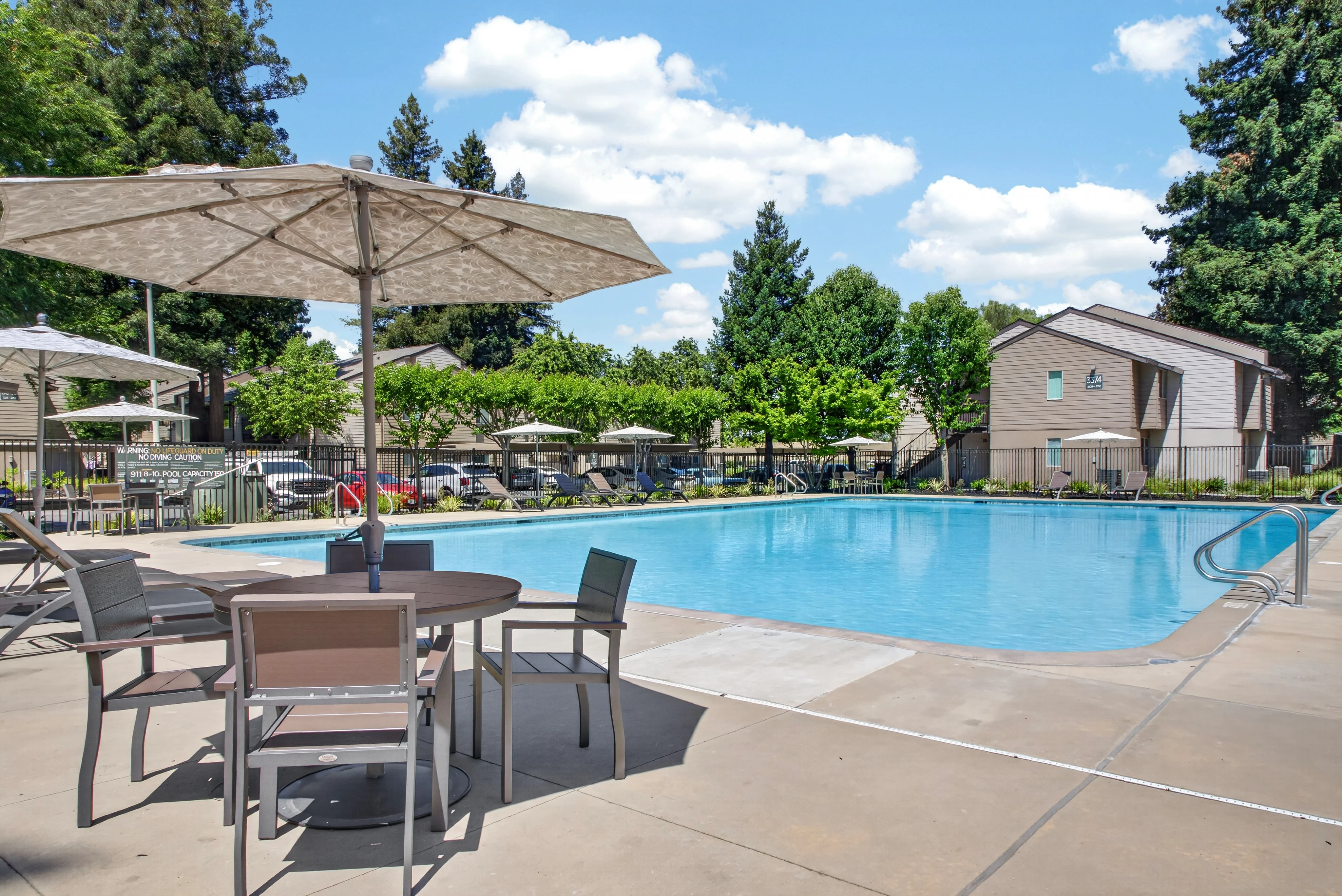 A well-maintained swimming pool area featuring a clear blue pool, surrounded by lounge chairs, umbrellas, and lush green trees under a bright blue sky with white clouds.