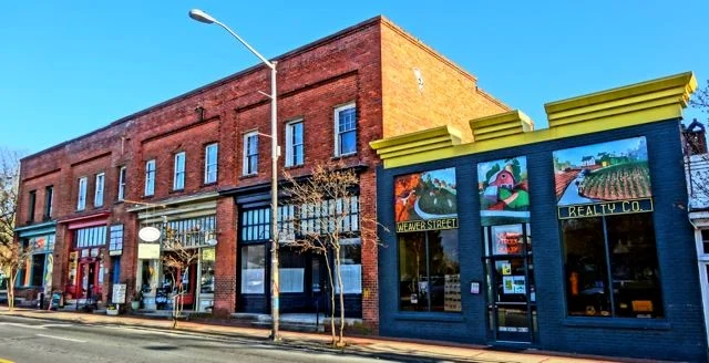 A row of historic buildings along a street, featuring brick facades and colorful storefronts. The sky is clear and blue.