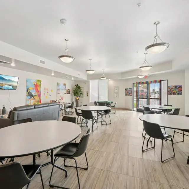 Community Room Overview A spacious community room with several round tables and chairs, natural light coming from large windows, and colorful art on the walls. A couch is present along with a television.
