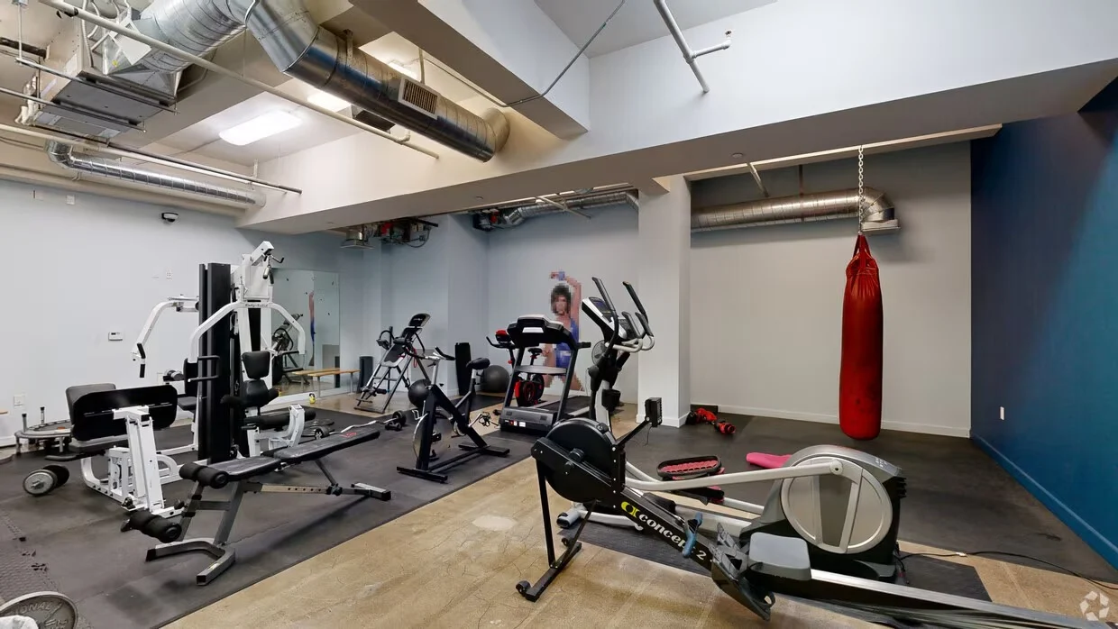 An interior view of a gym with various workout machines and equipment including an exercise bike, treadmill, and punching bag.