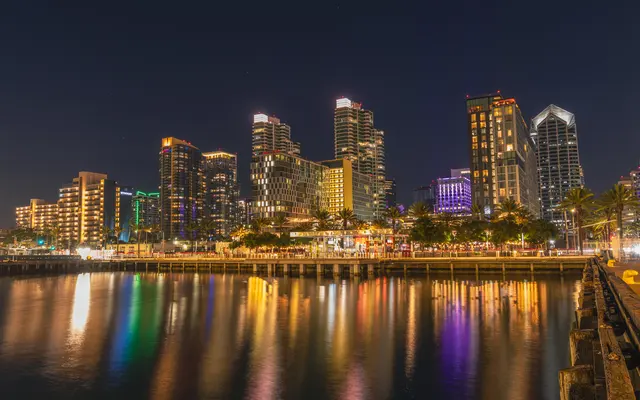A vibrant city skyline at night, showcasing tall buildings illuminated with colorful lights alongside a calm water body reflecting the lights.