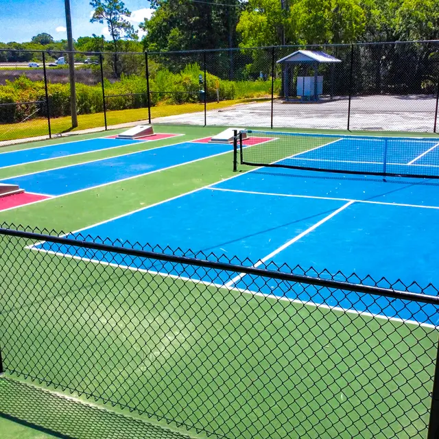 An aerial view of two tennis courts with vibrant blue surfaces, surrounded by a black chain-link fence. A small shelter is visible in the background, and the area is lush with greenery.