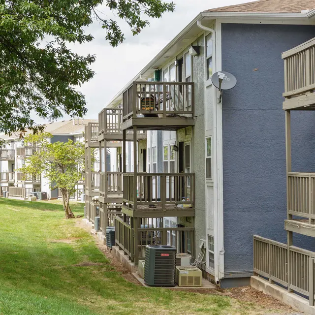 Exterior view of an apartment building with multiple balconies and air conditioning units, surrounded by green grass and trees.