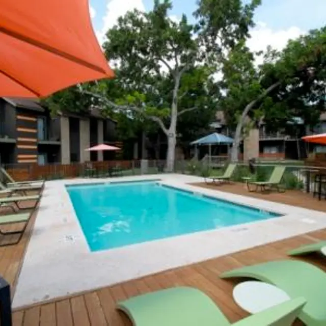 A modern apartment complex pool area featuring a rectangular swimming pool surrounded by lounge chairs and umbrellas. The pool area has wooden decking and is shaded by trees, with seating areas visible.
