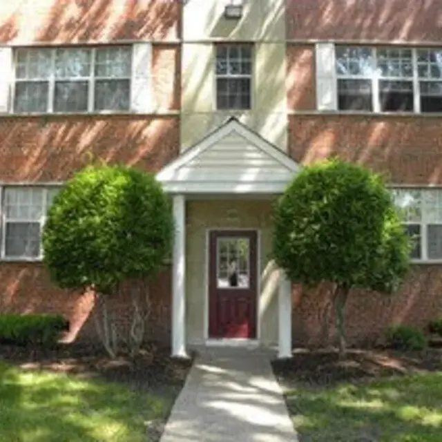 Front entrance of a brick apartment building with manicured bushes on either side of the pathway leading to the door.