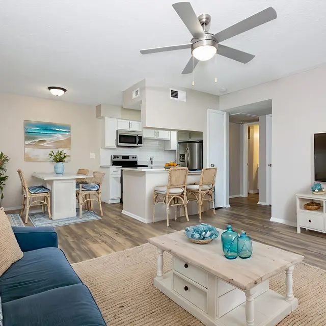 A modern living room featuring a blue sofa, a light coffee table, and a mounted flat-screen TV. In the background, there is a small dining area with bar stools and a kitchen with stainless steel appliances.