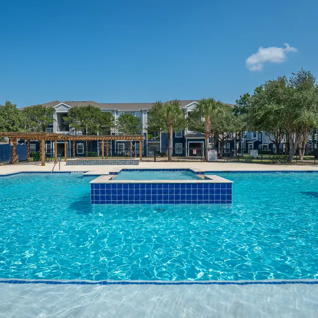 A large swimming pool featuring crystal clear water with a central raised platform. Lush greenery and residential buildings are visible in the background under a bright blue sky.