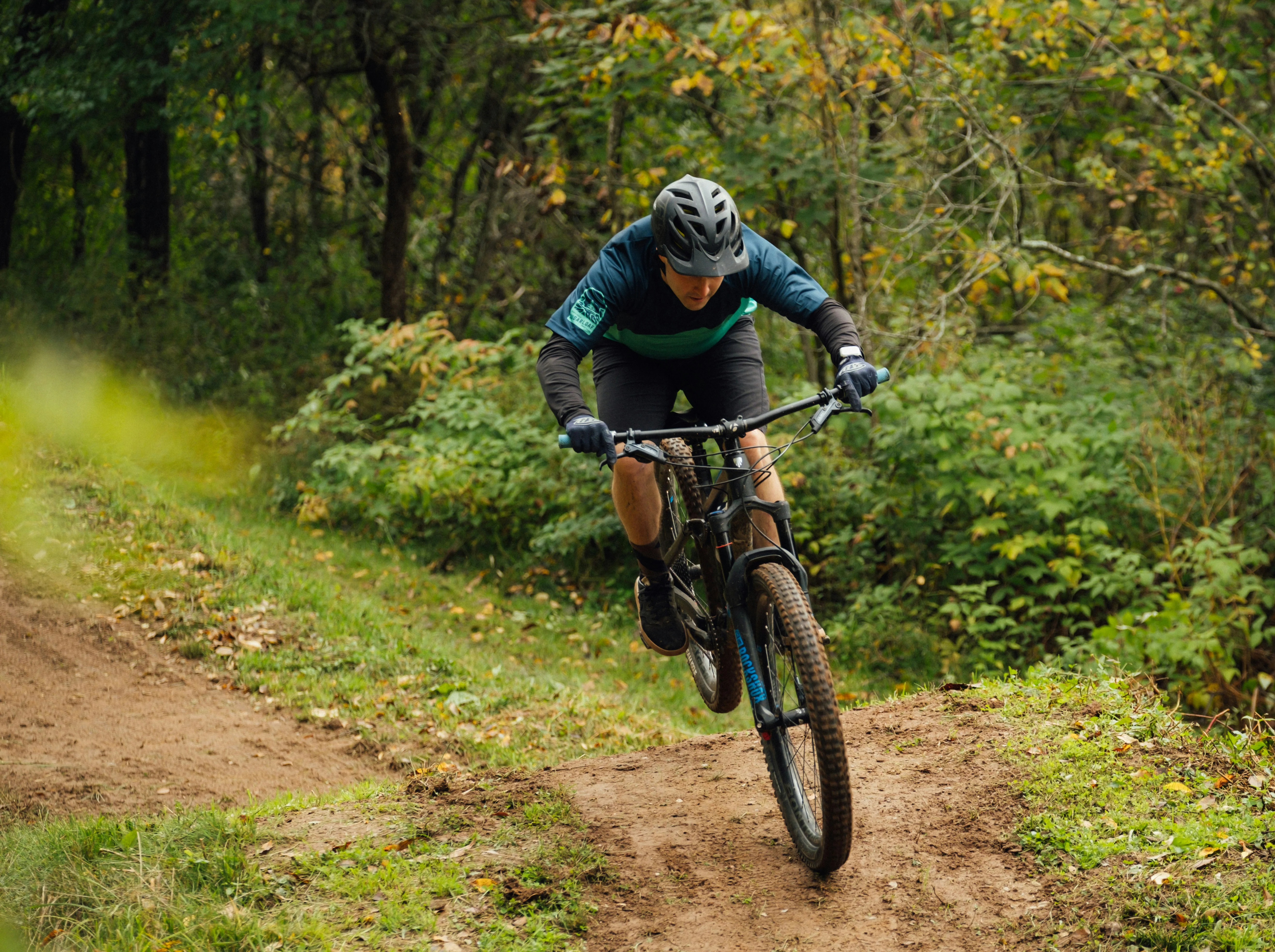 Mountain Biking Adventure A mountain biker in a blue and black outfit jumps off a dirt path surrounded by greenery.