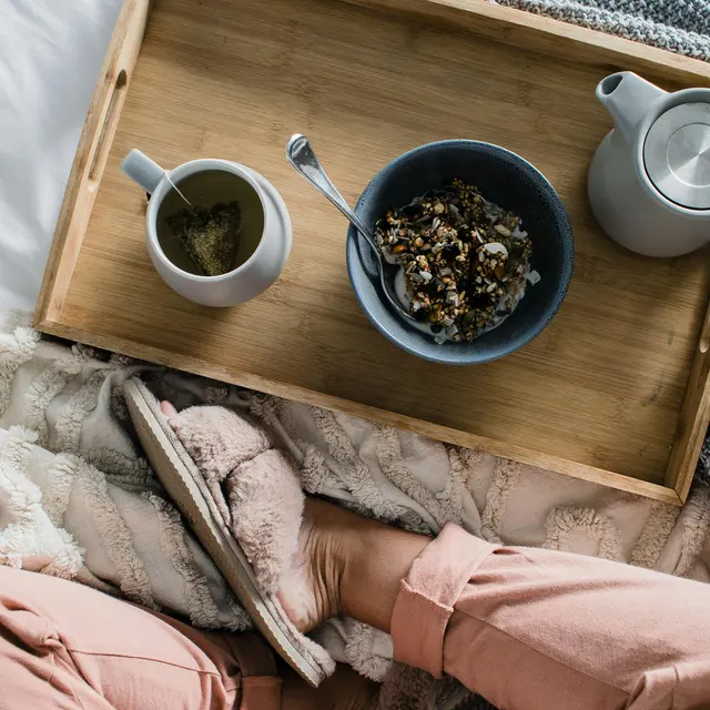 A cozy breakfast scene featuring a wooden tray with a bowl of granola, a cup of beverage, and a teapot, set on a soft blanket. A person's feet in cozy slippers are visible in the foreground.