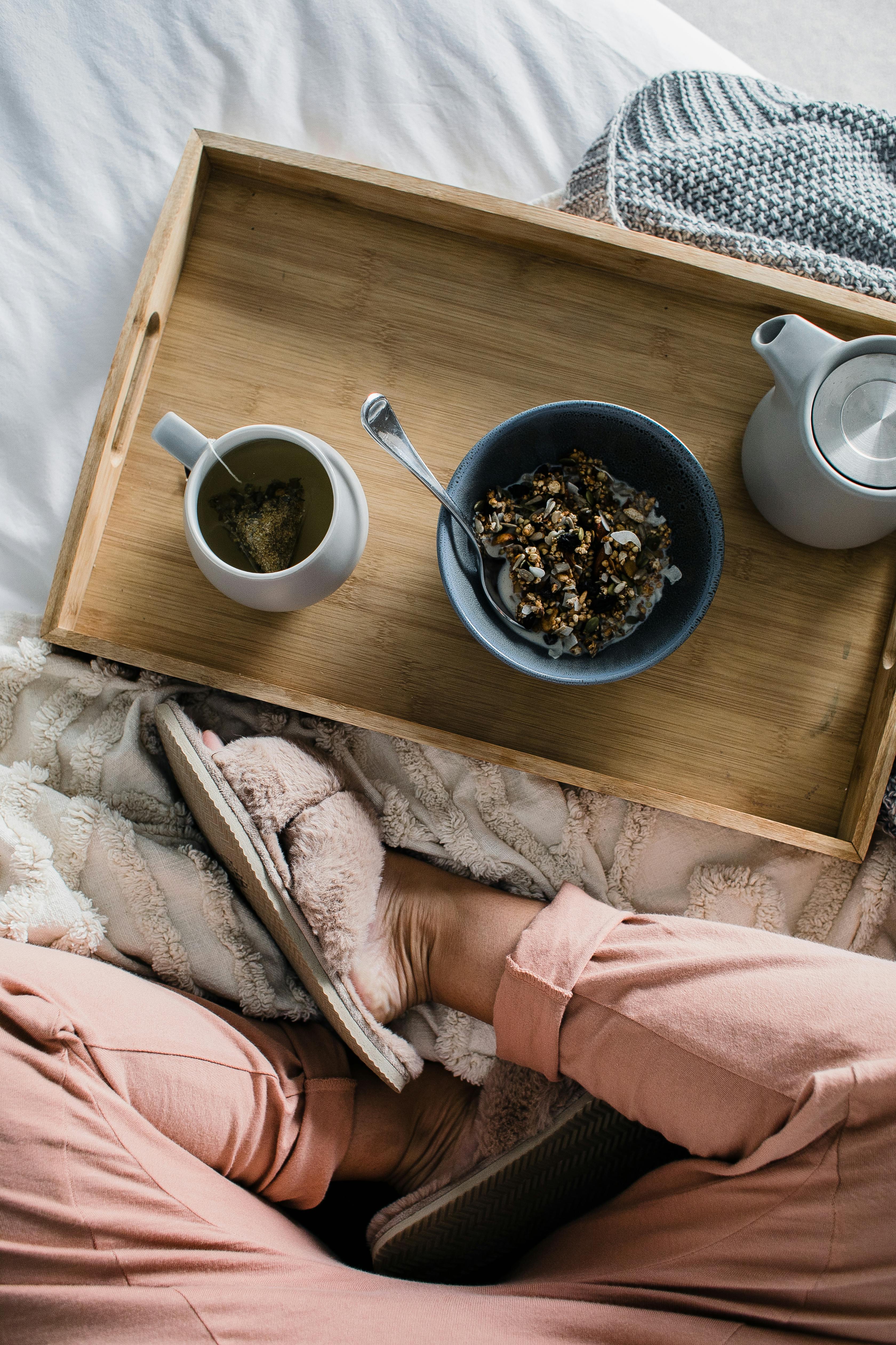A cozy breakfast scene featuring a wooden tray with a bowl of granola, a cup of beverage, and a teapot, set on a soft blanket. A person's feet in cozy slippers are visible in the foreground.