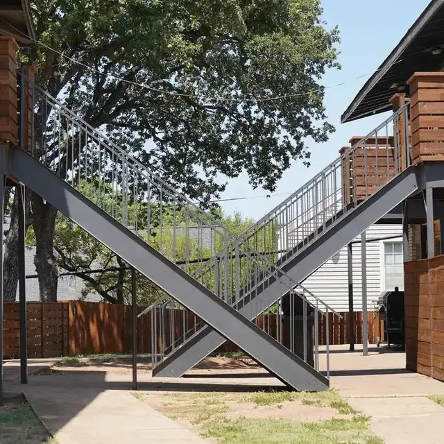 A set of outdoor metal stairs crossing in a zigzag pattern between two buildings, surrounded by trees and fenced areas.