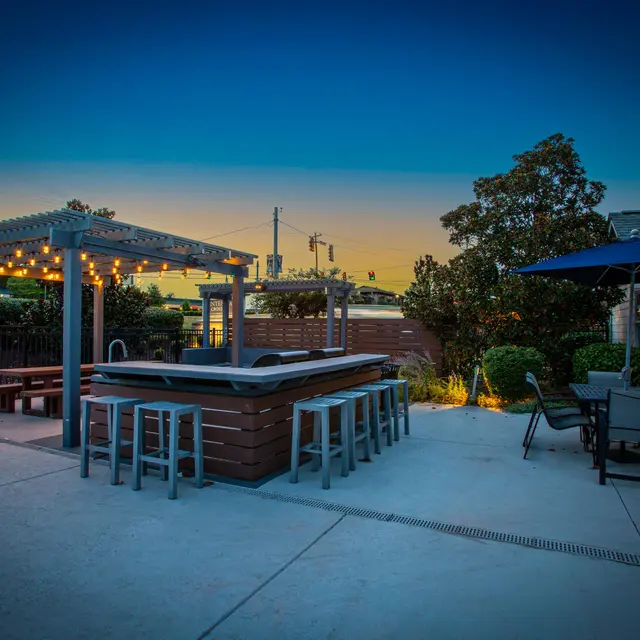 A stylish outdoor patio area featuring a bar with stools, a pergola adorned with string lights, and a seating arrangement, all under a twilight sky.