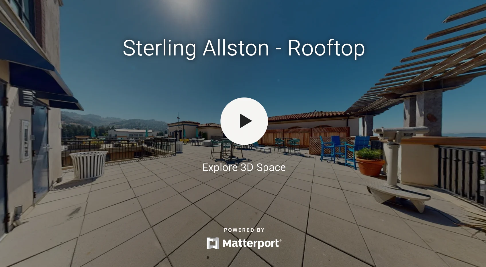 A panoramic view of a rooftop terrace featuring tiled flooring, blue lounge chairs, and scenic mountain views under a clear sky.
