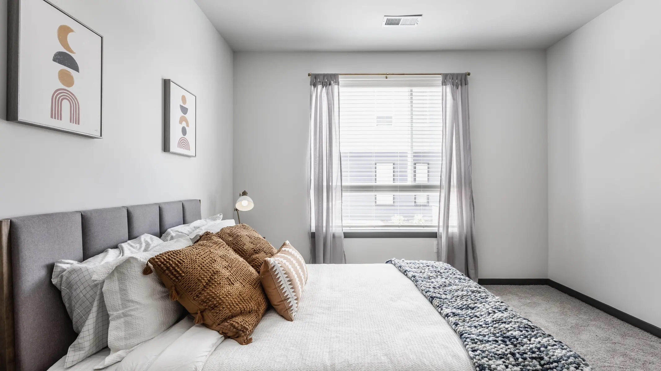 A modern bedroom with a neatly made bed featuring decorative pillows, large window with sheer curtains, and framed artwork on the walls.