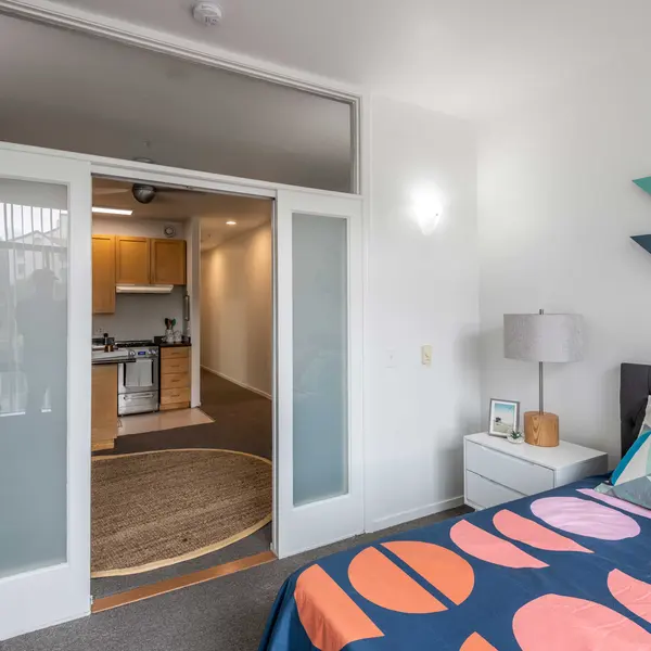 A stylish bedroom featuring a bed with a colorful duvet cover, nightstand, and a geometric wall decoration. A doorway leads into a kitchen area in the background.