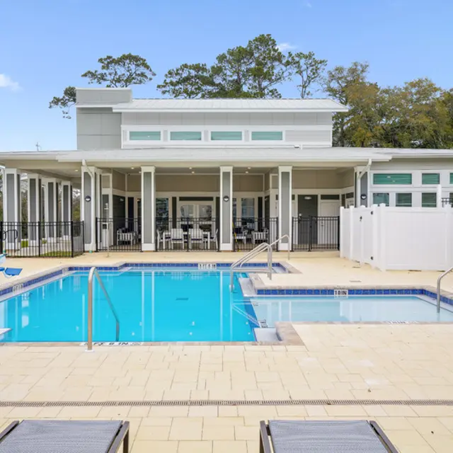 Modern pool area with lounge chairs, a swimming pool, and a clubhouse surrounded by trees under a clear blue sky.