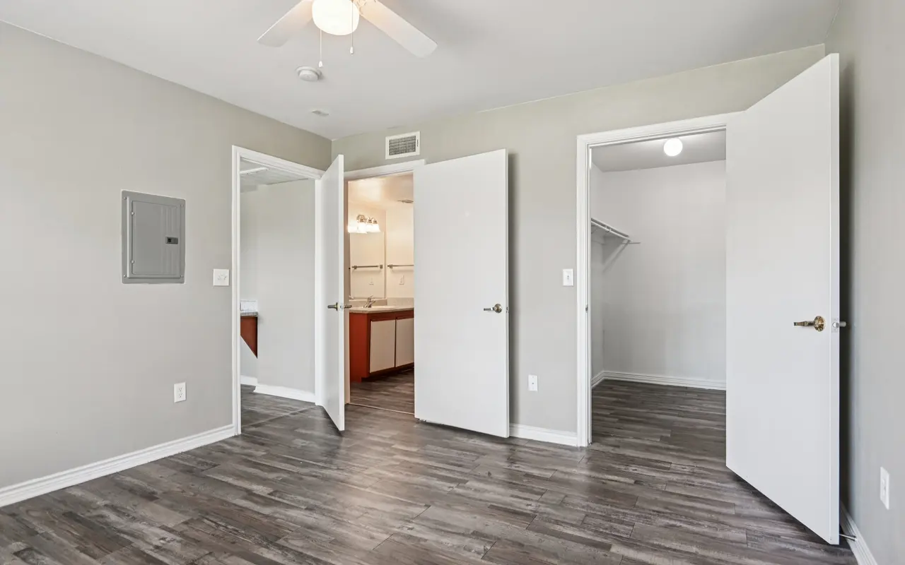 An empty room with a ceiling fan, light-colored walls, and dark wood flooring. There are three doors visible: one leading to a bathroom, another to a closet, and the third leading to what appears to be a kitchenette area.