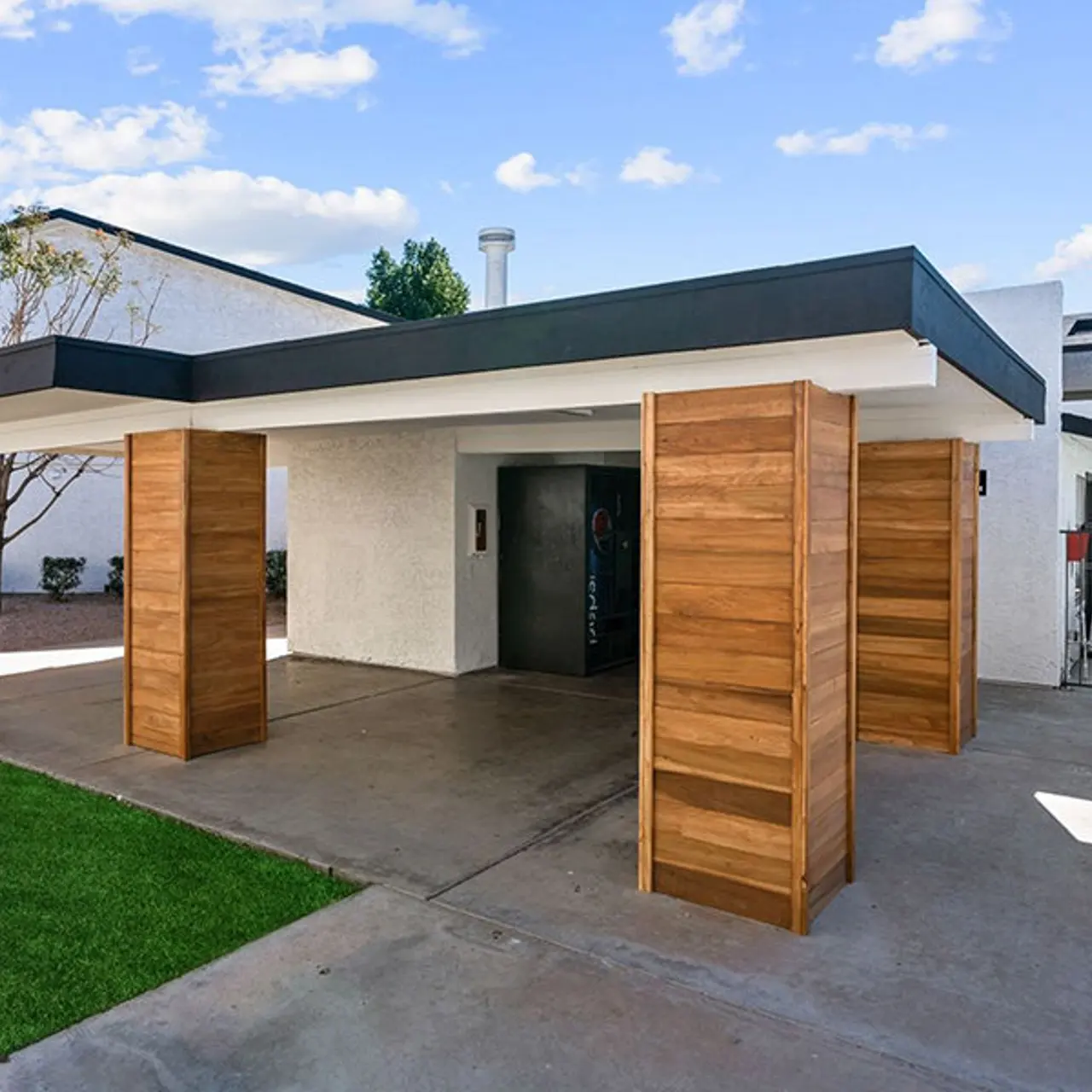 A modern entryway featuring a flat roof supported by wooden pillars, with a concrete floor and green lawn area in front. The background shows a fenced area with lounge chairs and structures behind the entryway.