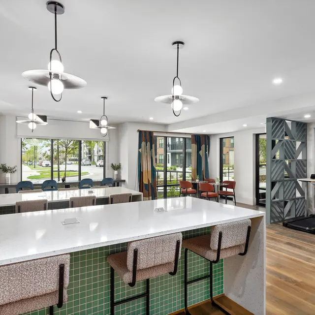 A stylish lounge area featuring a long white bar with green tile accents, bar stools, and large windows providing natural light. The space is decorated with modern light fixtures and colorful curtains, creating a warm and inviting atmosphere.