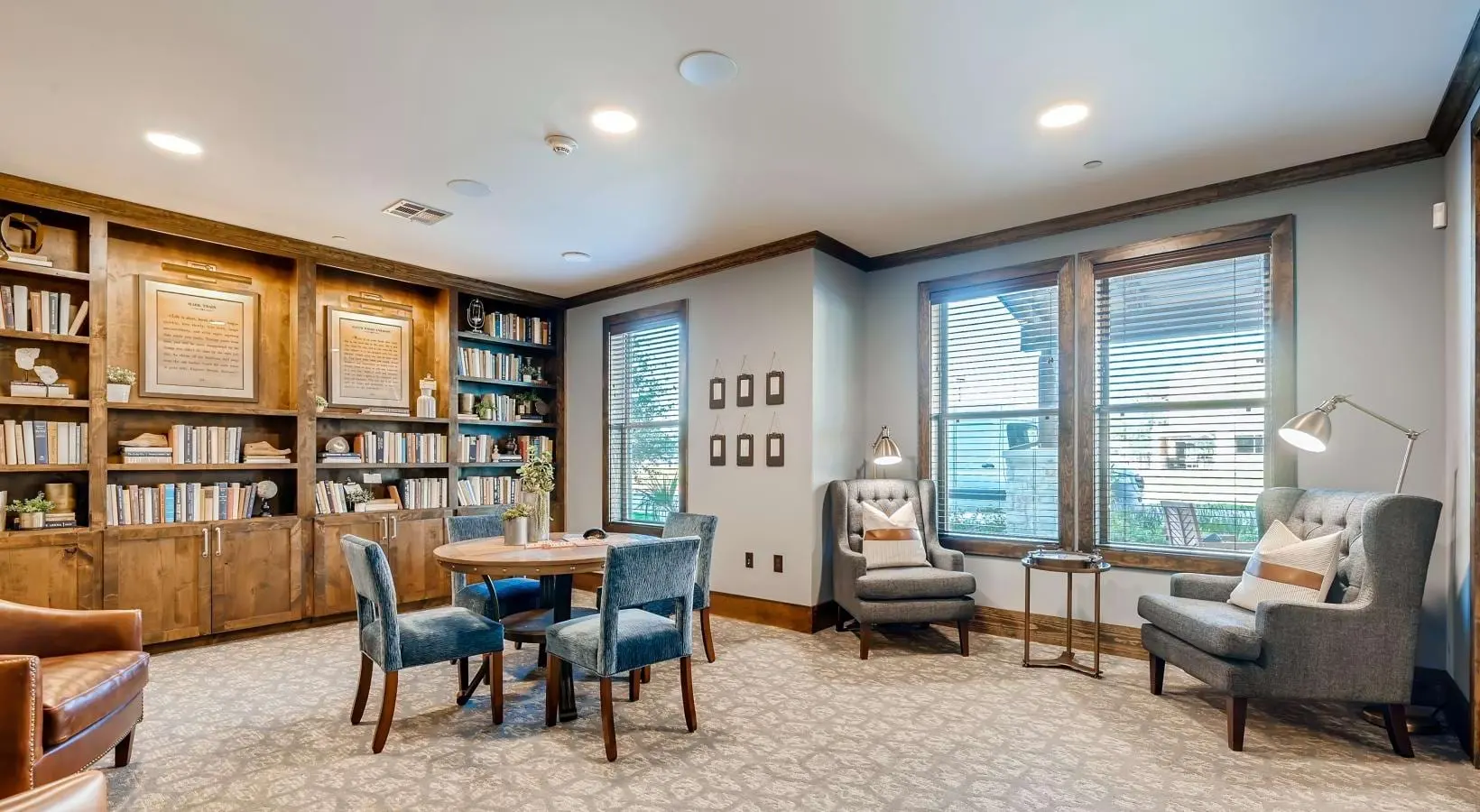 A cozy reading room featuring a wooden bookshelf filled with books, a small round table surrounded by blue chairs, and comfortable armchairs near large windows with natural light coming in.