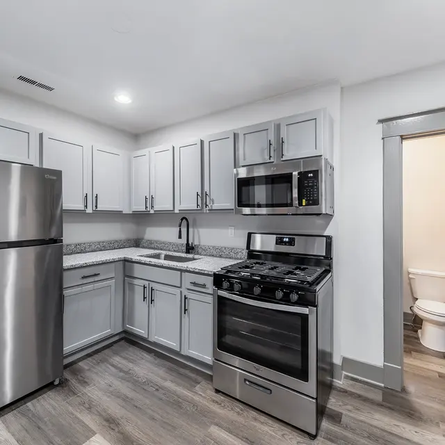 A modern kitchen featuring gray cabinetry, stainless steel appliances including a refrigerator and a stove, and a microwave. The flooring is a light wood finish, and there is a doorway leading to a bathroom with visible white fixtures.