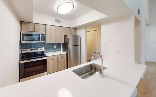 The Gleason A modern kitchen with wood cabinetry and stainless steel appliances. A sink is positioned on a white countertop with a view of the living space beyond.