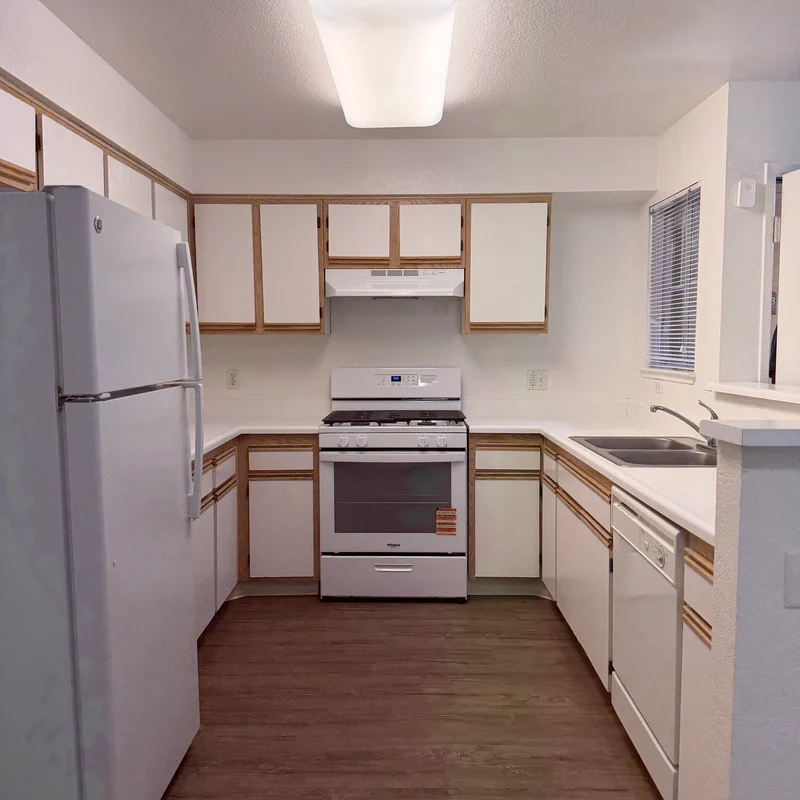 A clean, modern kitchen with white cabinets and appliances. The kitchen features a white refrigerator, a stove with an overhead exhaust, and a sink. The floor is laminate wood, creating a warm ambiance. Natural light comes through a window next to the sink.