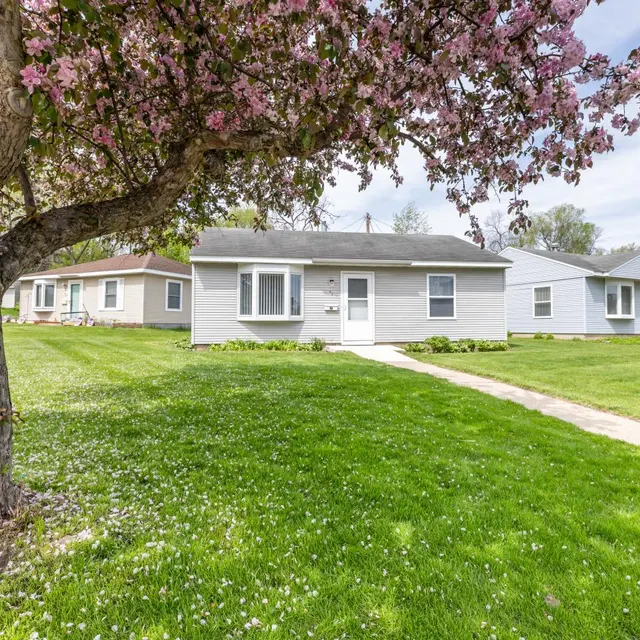 A suburban house surrounded by green grass and blooming trees. The house is light gray with a white front door, set on a lawn with a pathway leading up to it. Nearby houses are also visible.