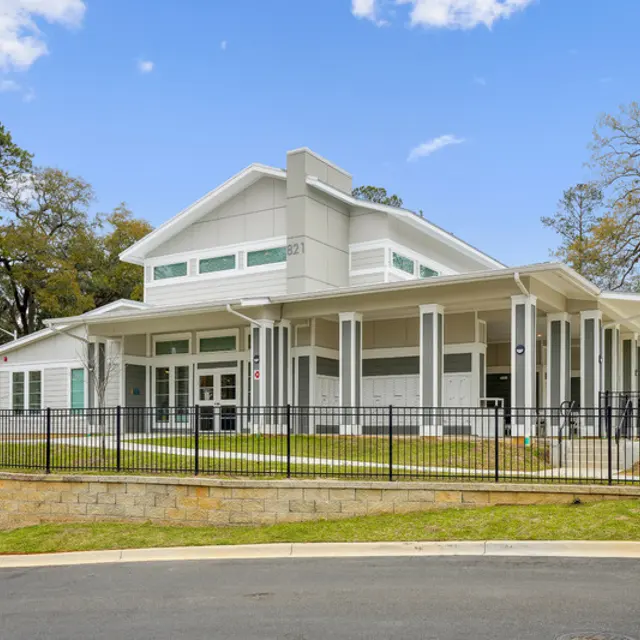 Modern building with a white exterior and large windows, surrounded by a green lawn and a black fence, under a blue sky.