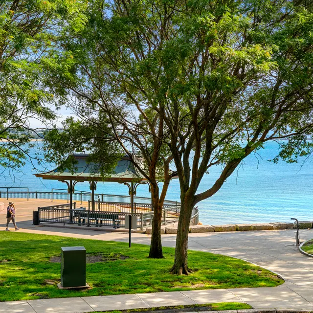 A waterfront park scene featuring green grass, trees, and a gazebo overlooking a calm lake. A path leads through the park with a person walking in the distance.