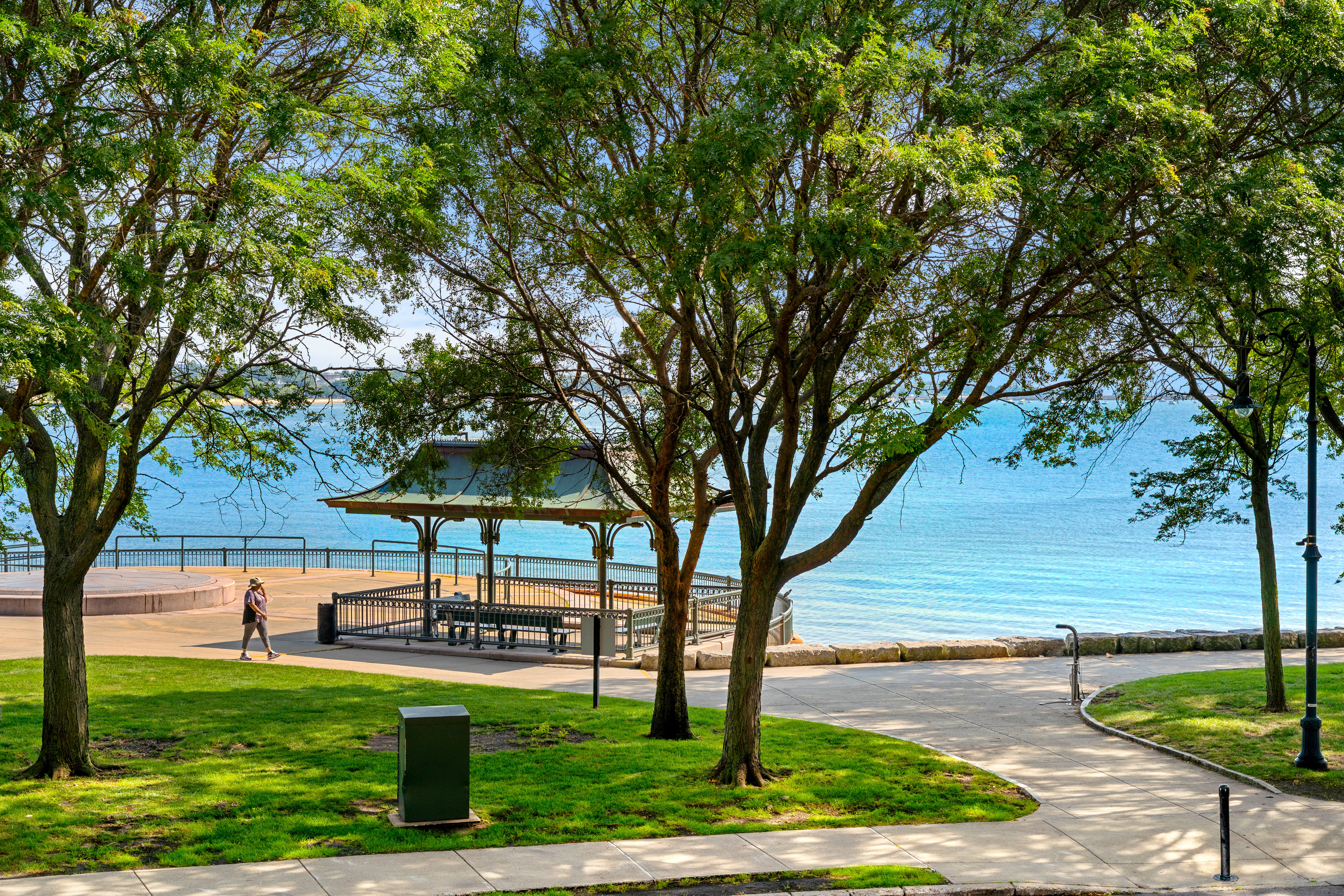 Serene Lakefront Park A waterfront park scene featuring green grass, trees, and a gazebo overlooking a calm lake. A path leads through the park with a person walking in the distance.