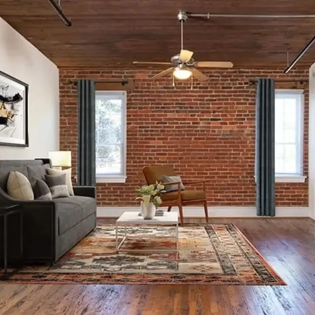 A modern living room featuring exposed brick walls, a brown ceiling fan, and large windows with sheer curtains. The space includes a gray sofa with throw pillows, a white coffee table on a patterned rug, and a brown armchair. A small media console is positioned against the wall, with a flat-screen television mounted nearby.