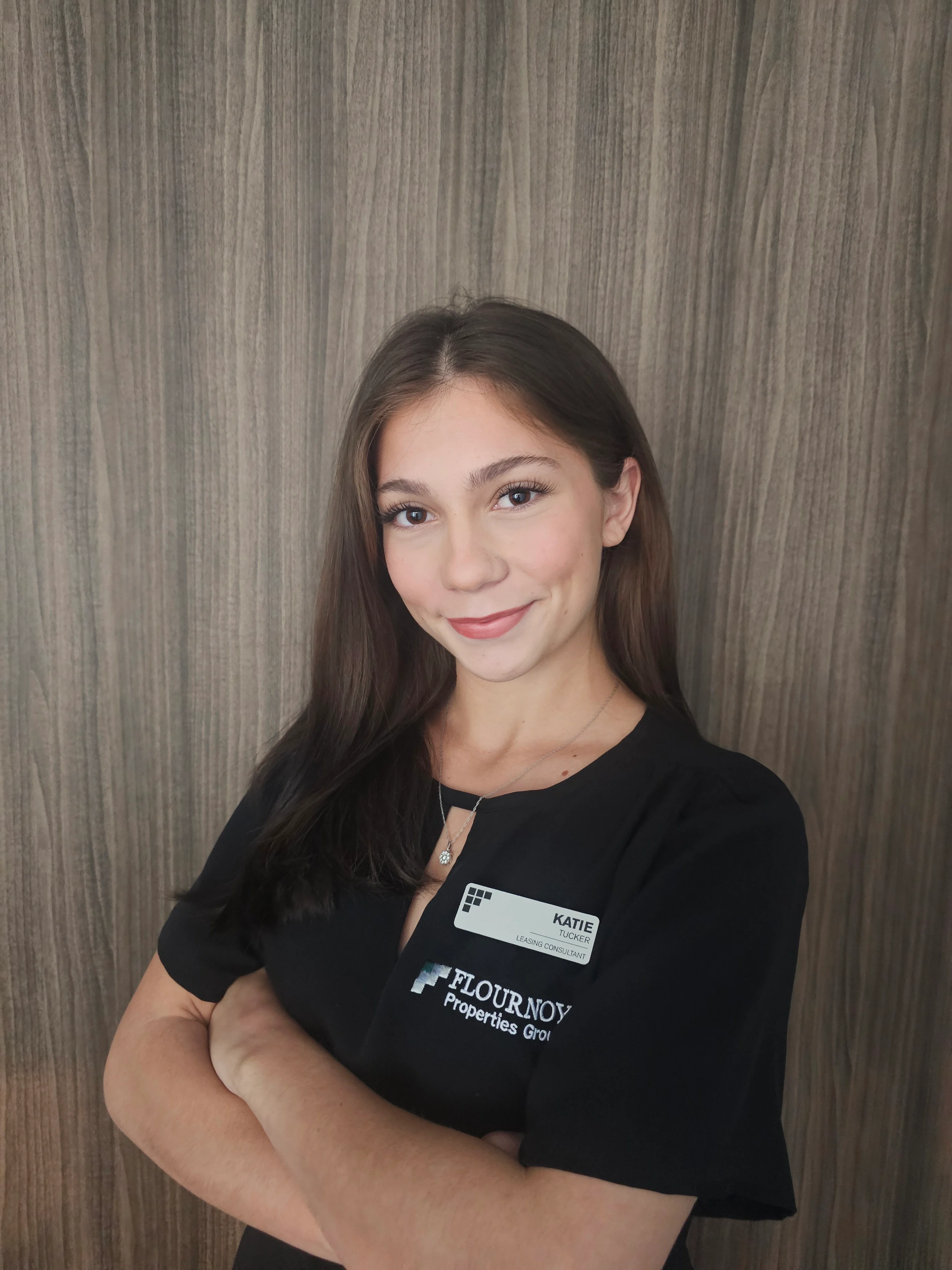Professional Portrait of a Woman A woman with long brown hair is standing with her arms crossed, wearing a black professional shirt with a name tag. The background is a wooden panel.